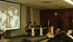 写真�@：右から代表の永井裕之さん、シンポジストの中医協委員の勝村久司さん、熊本大学医学部助教授の粂和彦さんスクリーン中央に写っているのは、事故で亡くなった永井さんの夫人